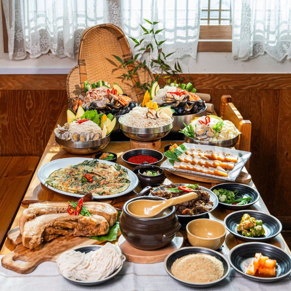 레이스 커튼 창가 나무 탁자에 차린 상 — 해물과 면을 담은 냄비, 파전, 수육, 반찬