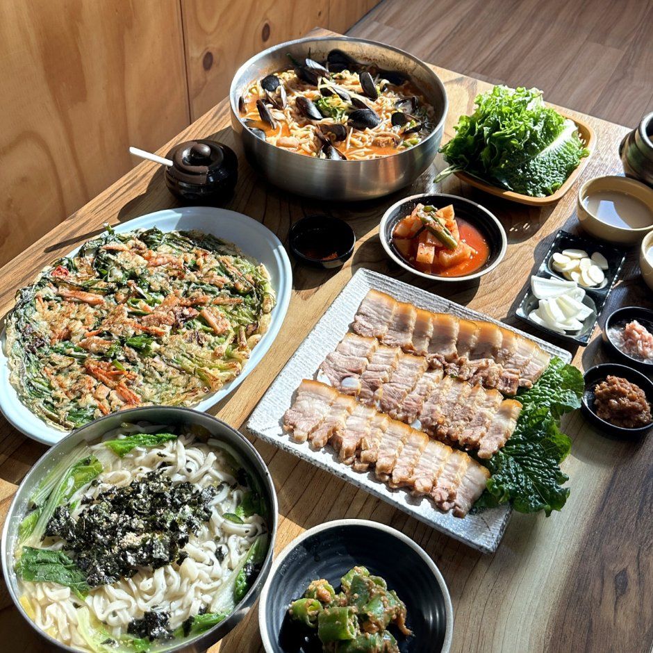 칼국수, 파전, 쌈 채소를 곁들인 수육, 홍합과 면이 든 붉은 국물 냄비와 반찬을 차린 상